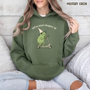 May include: Military green hoodie with a frog graphic riding a scooter and wearing a wizard hat. The text "Off to start chapter 40" is above the frog. The hoodie has a drawstring hood and a front pocket.
