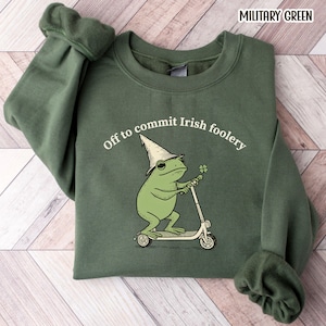 May include: Military green sweatshirt with the text "Off to commit Irish foolery." The graphic features a green frog wearing a hat and riding a scooter while holding a four-leaf clover. The sweatshirt has a crew neck and long sleeves.