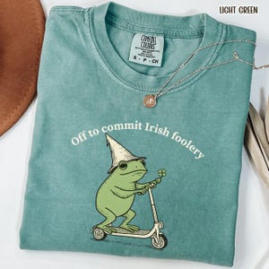 May include: Light green t-shirt with the text "Off to commit Irish foolery" and a graphic of a green frog wearing a hat and riding a scooter. The shirt also has a Comfort Colors tag.