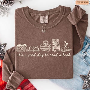 May include: A brown long-sleeve shirt with the phrase "it's a good day to read a book" in white. The graphic includes illustrations of books, a coffee cup, and hearts. A silver necklace with a sun pendant is visible.