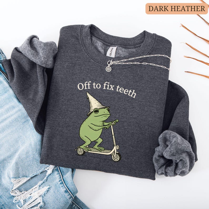 May include: Dark heather gray sweatshirt with the text "Off to fix teeth." The graphic features a green frog wearing a hat and riding a scooter. A silver necklace is draped across the sweatshirt.