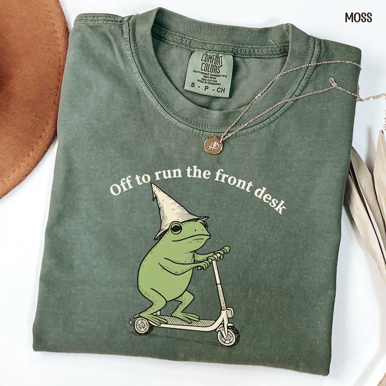 K&ouml;nnte beinhalten: Moosgr&uuml;nes T-Shirt mit dem Text "Off to run the front desk". Auf der Vorderseite ist ein Cartoon-Frosch mit Hut auf einem Roller abgebildet. Eine goldene Halskette liegt auf dem gefalteten Shirt.