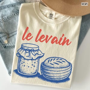 May include: Ivory-colored t-shirt with the words "le levain" in red script. Below the text is a blue and white illustration of a jar and a bowl. The shirt is folded on top of blue jeans and a tan hat.