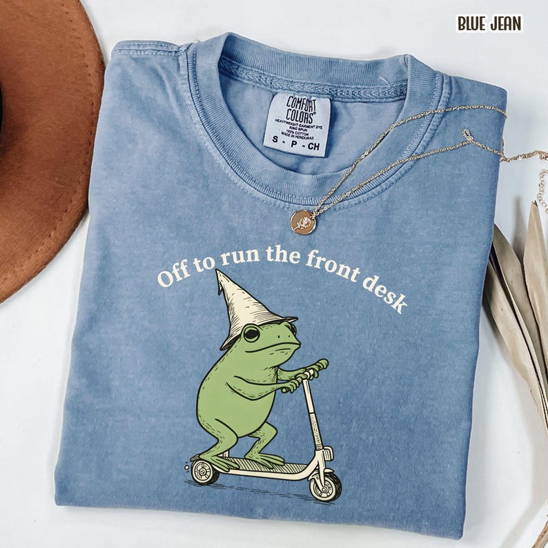 K&ouml;nnte beinhalten: Blaues Comfort Colors T-Shirt mit dem Text "Off to run the front desk". Das Shirt zeigt einen gr&uuml;nen Frosch mit Hut, der einen Roller f&auml;hrt. Eine goldene Halskette liegt &uuml;ber dem Shirt.