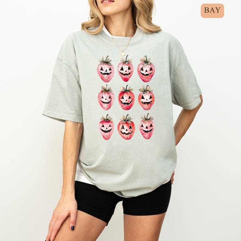 May include: A light green t-shirt featuring a repeating pattern of strawberry jack-o'-lanterns. The strawberries are red with green stems and carved faces. The shirt is a relaxed fit and has short sleeves.