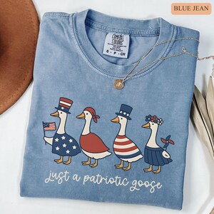 May include: Blue jean-colored t-shirt featuring a patriotic goose design. The graphic includes four geese dressed in red, white, and blue attire, with the text "just a patriotic goose" below the geese.