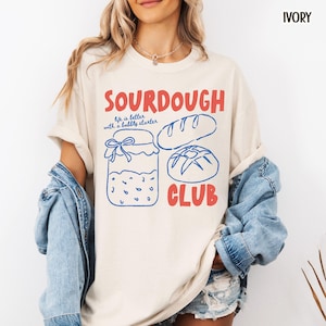 May include: Ivory-colored t-shirt with the words "SOURDOUGH CLUB" in red and blue. The shirt features a graphic of a jar, a loaf of bread, and a round loaf of bread. The text "life is better with a bubbly starter" is also included.