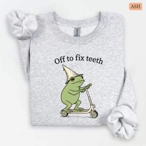 May include: Ash-colored sweatshirt with the text "Off to fix teeth." The graphic features a green frog wearing a hat and riding a scooter. The sweatshirt is made of a soft material and has long sleeves.