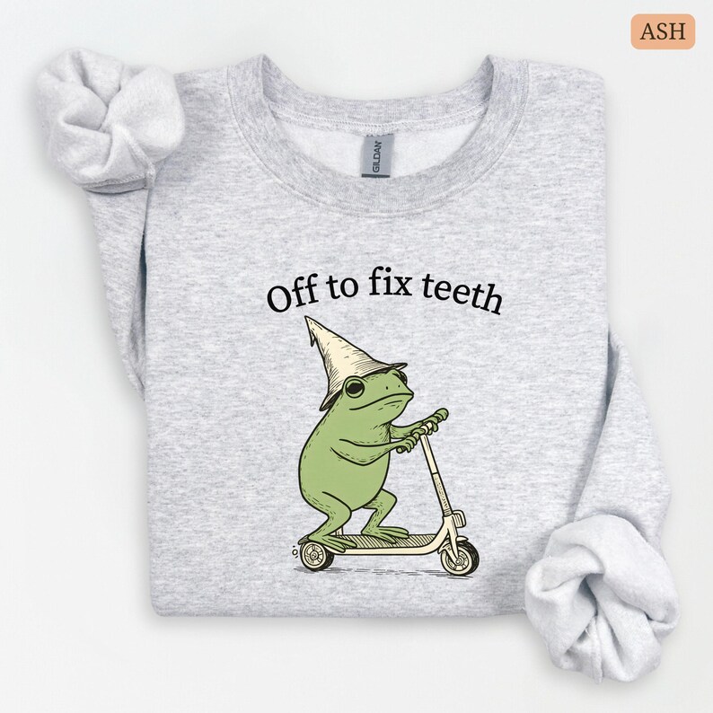 May include: Ash-colored sweatshirt with the text "Off to fix teeth." The graphic features a green frog wearing a hat and riding a scooter. The sweatshirt is made of a soft material and has long sleeves.
