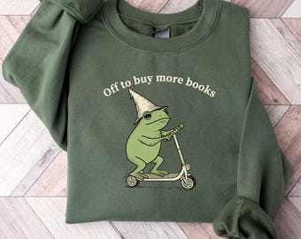 May include: Military green sweatshirt with the text "Off to buy more books." The graphic features a green frog wearing a wizard hat and riding a scooter. The sweatshirt is a solid color.