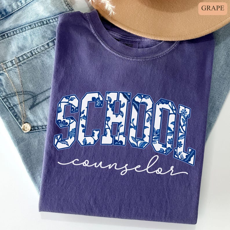 May include: A grape-colored t-shirt with the words "SCHOOL counselor" printed on the front. The word "SCHOOL" is in large, white block letters with a blue floral pattern. The word "counselor" is in a white cursive font.