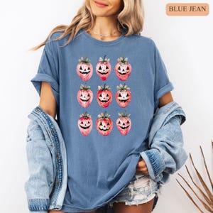 May include: Blue jean-colored t-shirt featuring a repeating pattern of strawberries carved like jack-o'-lanterns. The strawberries are red and pink with green leaves. The text "BLUE JEAN" is in the upper right corner.