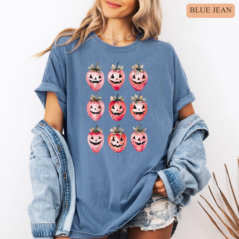 May include: Blue jean-colored t-shirt featuring a repeating pattern of strawberries carved like jack-o'-lanterns. The strawberries are red and pink with green leaves. The text "BLUE JEAN" is in the upper right corner.