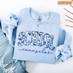 May include: Light blue sweatshirt with the words "SCHOOL counselor" in a floral design. The sweatshirt is folded on top of a pair of blue jeans. A silver necklace with a small pendant is on the sweatshirt.