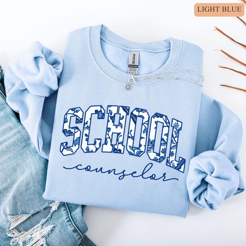 May include: Light blue sweatshirt with the words "SCHOOL counselor" in a floral design. The sweatshirt is folded on top of a pair of blue jeans. A silver necklace with a small pendant is on the sweatshirt.