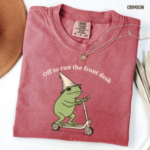 K&ouml;nnte beinhalten: Karmesinrotes T-Shirt mit einer Grafik einer gr&uuml;nen Froschfigur, die einen Zaubererhut tr&auml;gt und einen Roller f&auml;hrt. Der Text "Off to run the front desk" ist aufgedruckt. Eine silberne Halskette mit Anh&auml;nger ist zu sehen.