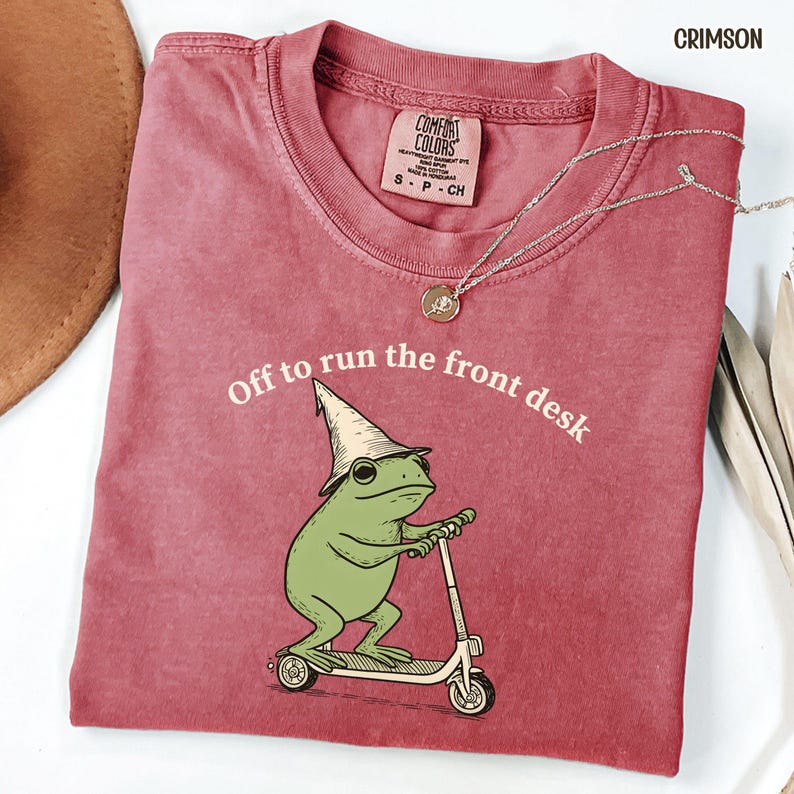 K&ouml;nnte beinhalten: Karmesinrotes T-Shirt mit einer Grafik einer gr&uuml;nen Froschfigur, die einen Zaubererhut tr&auml;gt und einen Roller f&auml;hrt. Der Text "Off to run the front desk" ist aufgedruckt. Eine silberne Halskette mit Anh&auml;nger ist zu sehen.