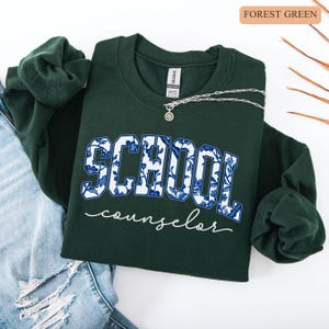 May include: Forest green sweatshirt with the words "SCHOOL counselor" in white and blue floral print. A silver necklace with a pendant is draped across the sweatshirt. The sweatshirt is folded on top of a pair of blue jeans.