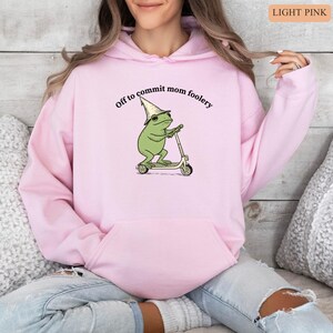 May include: Light pink hoodie with a front graphic of a green frog wearing a hat and riding a scooter. The text "Off to commit mom foolery" is above the frog. The hoodie has a kangaroo pocket and a drawstring hood.