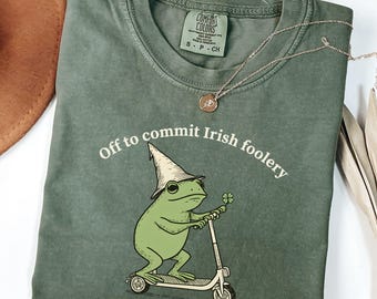 May include: Moss green t-shirt with the phrase "Off to commit Irish foolery" and a cartoon frog wearing a hat and riding a scooter. The frog is holding a four-leaf clover. A gold necklace is draped on the shirt.