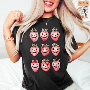 May include: Black t-shirt featuring a repeating pattern of strawberry-shaped jack-o'-lanterns. The strawberries are pink with green stems and leaves, and each has a carved jack-o'-lantern face. The word "BLACK" is in the upper right corner.