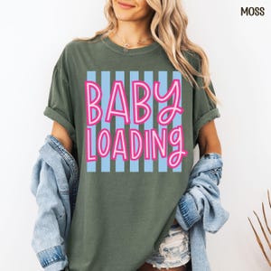 May include: Moss green t-shirt with the words "BABY LOADING" in pink lettering over a light blue striped background. The shirt has rolled-up sleeves and is paired with a denim jacket.