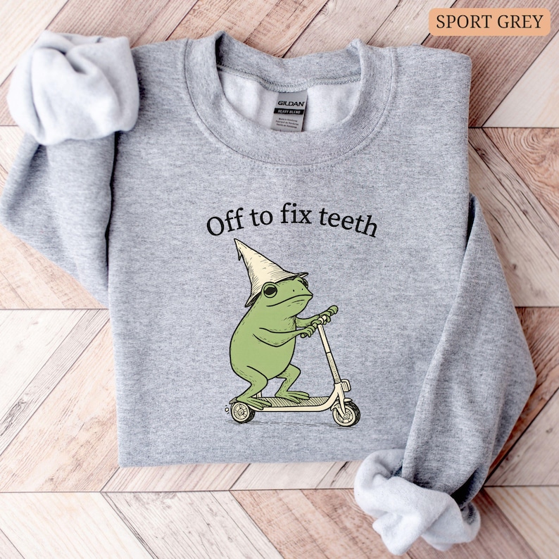 May include: Sport grey sweatshirt with the text "Off to fix teeth" above a cartoon frog riding a scooter. The frog is wearing a hat and glasses. The sweatshirt is a light gray color.
