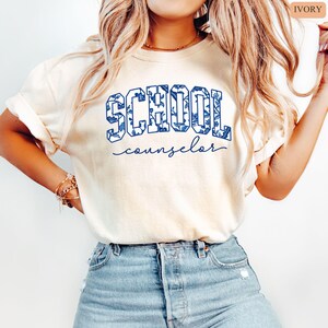 May include: Ivory-colored t-shirt with the word "SCHOOL" in blue floral print letters and the word "counselor" in a cursive font. The shirt is paired with blue jeans.