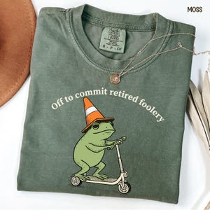 May include: A moss green t-shirt featuring a graphic of a green frog wearing an orange traffic cone and riding a scooter. The text above the frog reads, Off to commit retired foolery. The shirt has a crew neckline and a Comfort Colors brand label.