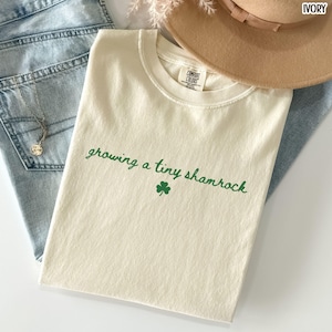 May include: Ivory-colored t-shirt with the text "growing a tiny shamrock" in green script, along with a green shamrock graphic. The shirt is laid on top of blue jeans and a beige hat.