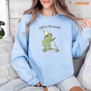 May include: Light blue sweatshirt featuring a cartoon frog riding a scooter, wearing a wizard hat and sunglasses. The text "Off to fix teeth" is above the frog. The sweatshirt is a solid light blue color.