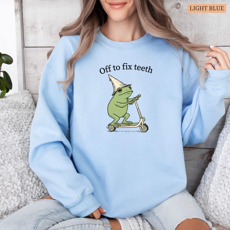 May include: Light blue sweatshirt featuring a cartoon frog riding a scooter, wearing a wizard hat and sunglasses. The text "Off to fix teeth" is above the frog. The sweatshirt is a solid light blue color.