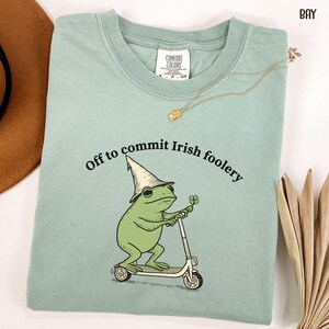 May include: A light green Comfort Colors t-shirt with a graphic of a green frog wearing a hat and riding a scooter, holding a four-leaf clover. The text "Off to commit Irish foolery" is arched above the frog.