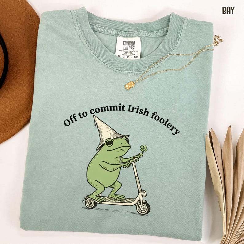 May include: A light green Comfort Colors t-shirt with a graphic of a green frog wearing a hat and riding a scooter, holding a four-leaf clover. The text "Off to commit Irish foolery" is arched above the frog.