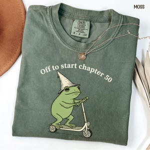 May include: Moss green t-shirt with the text "Off to start chapter 50" and a cartoon frog wearing a wizard hat and riding a scooter. A silver necklace with a pendant is on the shirt. The shirt is folded.