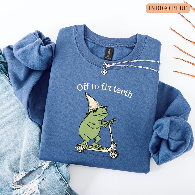 May include: Indigo blue sweatshirt with the text "Off to fix teeth." The graphic features a green frog wearing a wizard hat and sunglasses, riding a scooter. A silver necklace is draped on the sweatshirt.