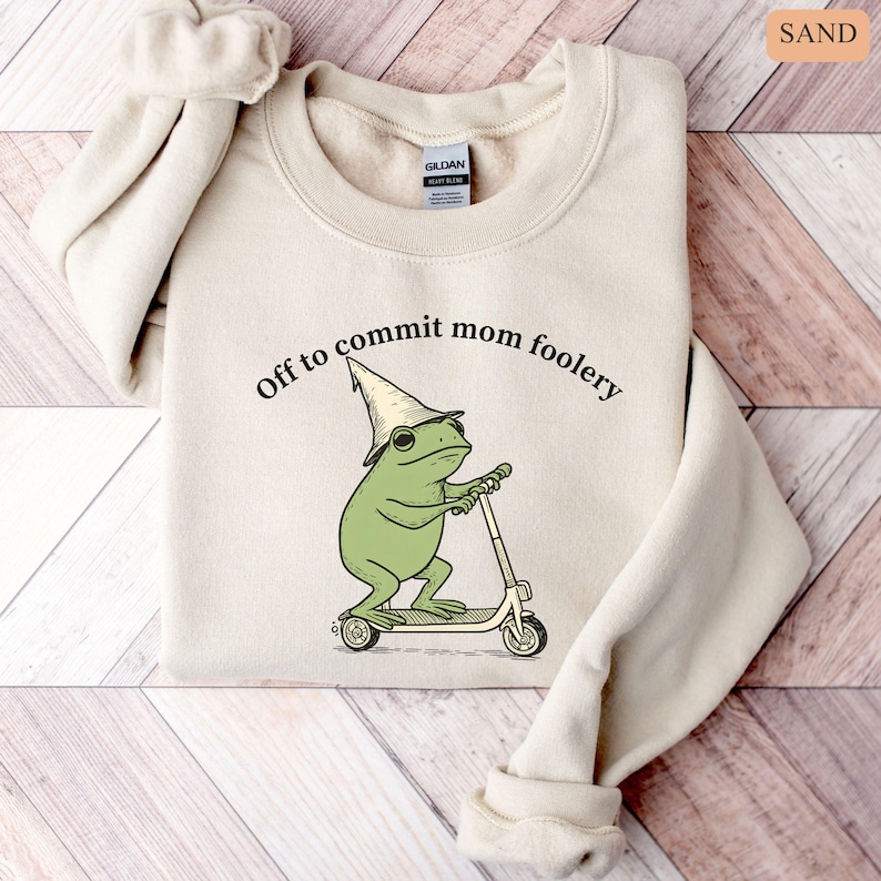 May include: Sand-colored sweatshirt with the text "Off to commit mom foolery." The graphic features a green frog wearing a wizard hat and riding a scooter. The sweatshirt is made of a soft material.