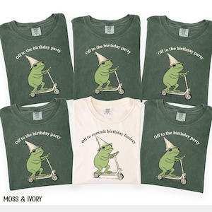 May include: A collection of green and cream-colored t-shirts. Each shirt features a graphic of a green frog on a scooter wearing a party hat, with the text "Off to the birthday party" or "Off to commit birthday foolery". The brand name "MOSS & IVORY" is at the bottom.