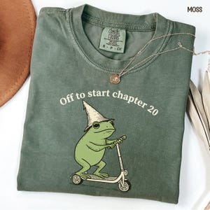 May include: Moss green t-shirt with the text "Off to start chapter 20" and a cartoon frog wearing a wizard hat and riding a scooter. A silver necklace with a pendant is on the shirt.