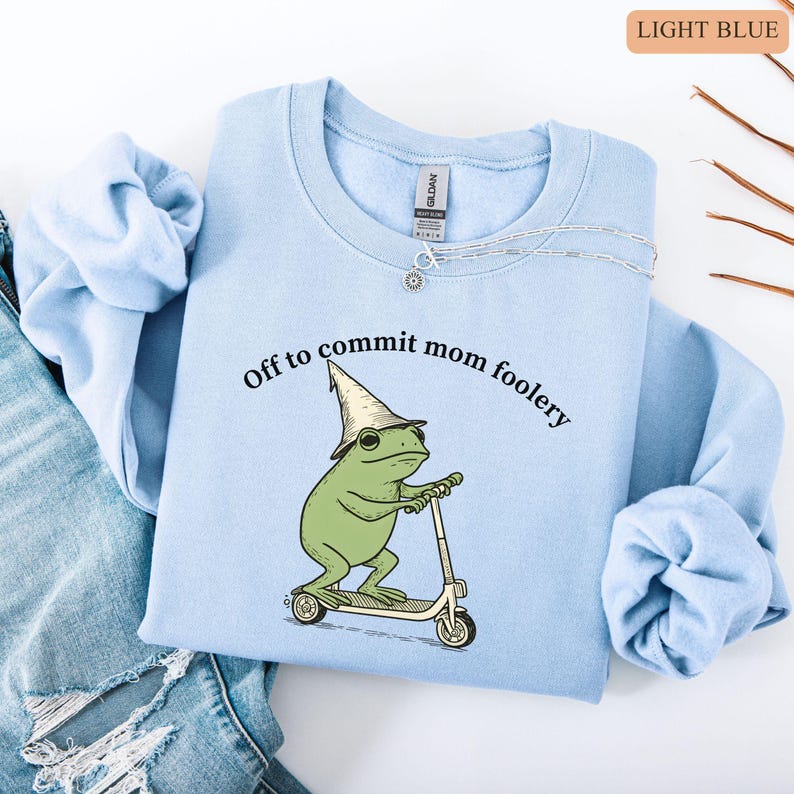 May include: Light blue sweatshirt with the text "Off to commit mom foolery" and a cartoon frog riding a scooter. The frog wears a wizard hat. A silver necklace is on the sweatshirt.