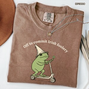 May include: Espresso-colored t-shirt with the text "Off to commit Irish foolery." The graphic features a green frog wearing a leprechaun hat and riding a scooter, holding a four-leaf clover. A silver necklace is draped across the shirt.