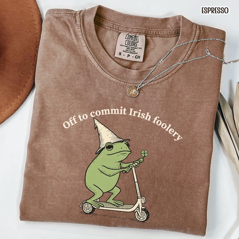 May include: Espresso-colored t-shirt with the text "Off to commit Irish foolery." The graphic features a green frog wearing a leprechaun hat and riding a scooter, holding a four-leaf clover. A silver necklace is draped across the shirt.