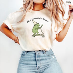 May include: Ivory-colored t-shirt featuring a green frog riding a scooter and wearing a hat. The frog holds a four-leaf clover. Text above the frog reads "Off to commit Irish foolery."