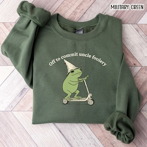 May include: Military green sweatshirt with the text "Off to commit uncle foolery." The graphic features a green frog wearing a wizard hat and riding a scooter. The sweatshirt has a crew neck and long sleeves.