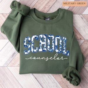 May include: Military green sweatshirt with the word "SCHOOL" in large, blue floral print letters and the word "counselor" in a cursive font. The sweatshirt is a solid color with ribbed cuffs.