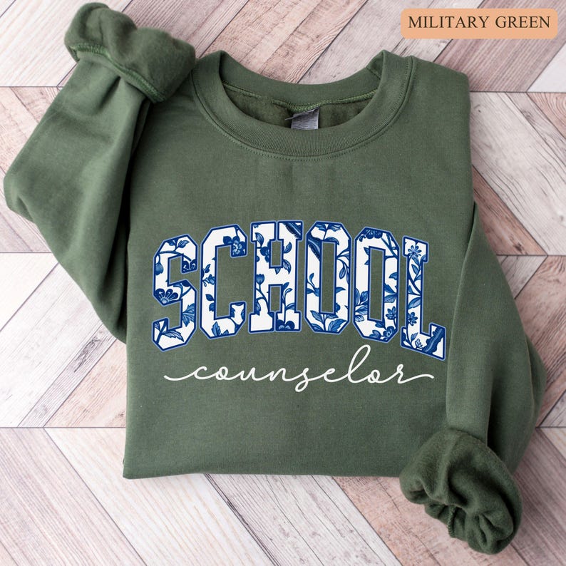May include: Military green sweatshirt with the word "SCHOOL" in large, blue floral print letters and the word "counselor" in a cursive font. The sweatshirt is a solid color with ribbed cuffs.