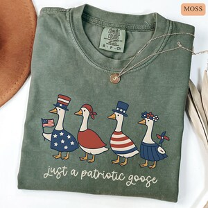 May include: Moss green Comfort Colors t-shirt featuring a patriotic goose design. The graphic shows four geese dressed in red, white, and blue attire, with the text "just a patriotic goose" below. A gold necklace is draped across the shirt.