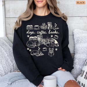 May include: Black sweatshirt with a cream-colored graphic featuring dogs, coffee cups, books, and paw prints. The text "dogs. coffee. books." is printed above the illustrations. The design includes sleeping dogs, stacks of books, and coffee cups.