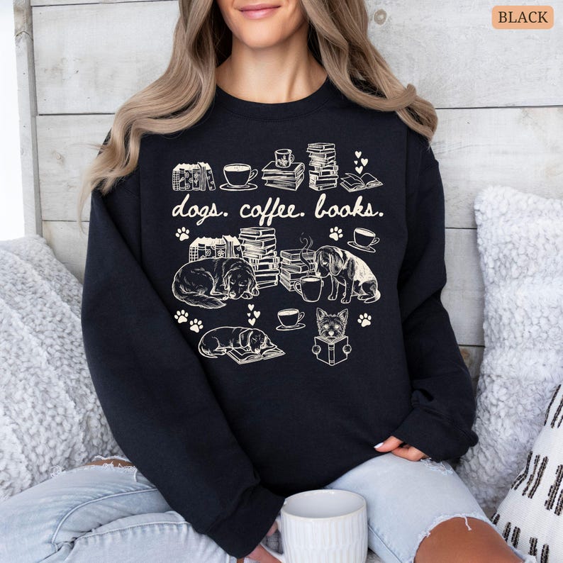 May include: Black sweatshirt with a cream-colored graphic featuring dogs, coffee cups, books, and paw prints. The text "dogs. coffee. books." is printed above the illustrations. The design includes sleeping dogs, stacks of books, and coffee cups.