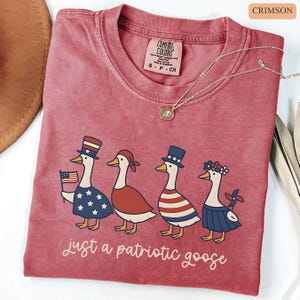 May include: Crimson-colored t-shirt featuring a patriotic goose design. The graphic shows four geese dressed in patriotic attire, including a hat, flag, and star-spangled clothing. The text "just a patriotic goose" is printed below the geese.